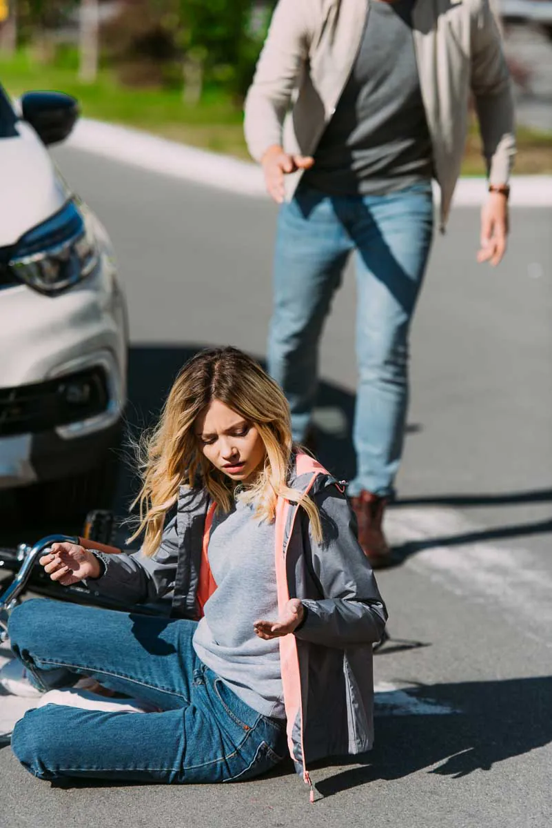 A distressed woman sits on the ground at a crosswalk next to a white car and a fallen bicycle as a man approaches to help after a road accident.